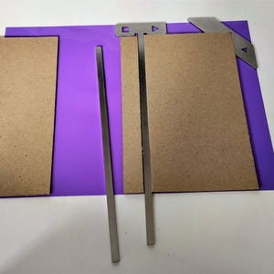 Può includere: Layout di un progetto artigianale con un tappetino viola, due pezzi di cartone marrone e diversi componenti metallici argentati. I componenti includono due barre lunghe e sottili e due pezzi angolati, il tutto disposto su una superficie bianca.