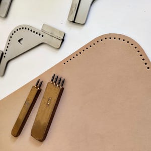 Peut inclure: Des outils de maroquinerie, dont des guides de couture en m&eacute;tal et des poin&ccedil;ons en laiton, dispos&eacute;s sur une pi&egrave;ce de cuir beige clair. Les outils sont con&ccedil;us pour cr&eacute;er des trous de couture espac&eacute;s uniform&eacute;ment.