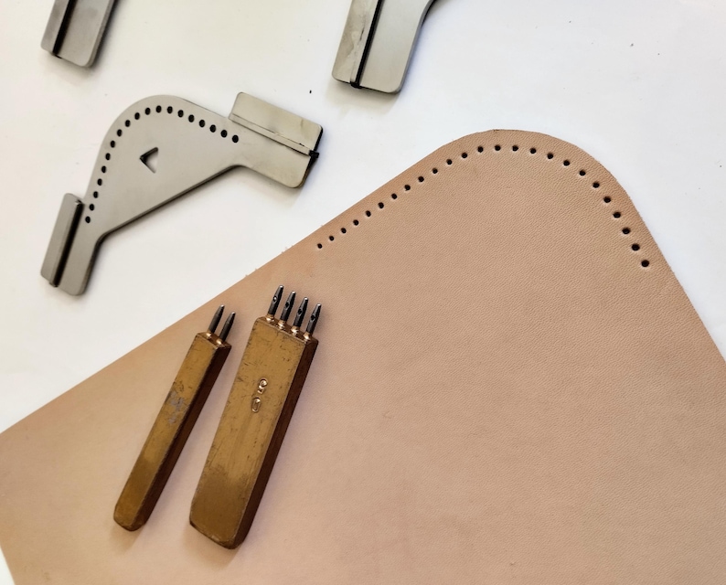 Peut inclure: Des outils de maroquinerie, dont des guides de couture en m&eacute;tal et des poin&ccedil;ons en laiton, dispos&eacute;s sur une pi&egrave;ce de cuir beige clair. Les outils sont con&ccedil;us pour cr&eacute;er des trous de couture espac&eacute;s uniform&eacute;ment.