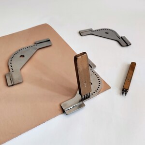 Peut inclure: Outils de travail du cuir, dont un rainureur &agrave; couture en m&eacute;tal, un poin&ccedil;on &agrave; cuir et un ciseau &agrave; couture &agrave; manche en bois. Les outils sont dispos&eacute;s sur une pi&egrave;ce de cuir brun clair, sur fond blanc.