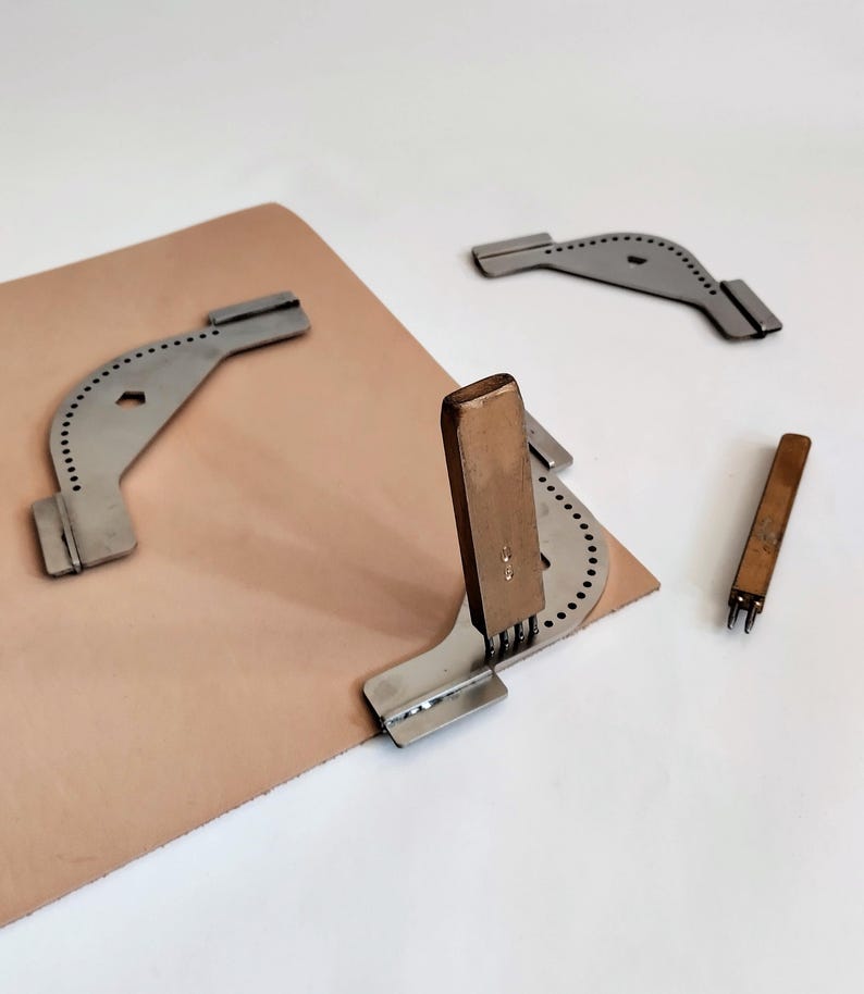 Peut inclure: Outils de travail du cuir, dont un rainureur &agrave; couture en m&eacute;tal, un poin&ccedil;on &agrave; cuir et un ciseau &agrave; couture &agrave; manche en bois. Les outils sont dispos&eacute;s sur une pi&egrave;ce de cuir brun clair, sur fond blanc.