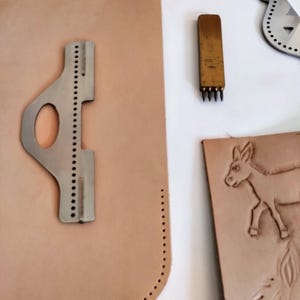 Peut inclure: Ensemble d'outils et de mat&eacute;riaux de maroquinerie. Comprend un guide de couture en m&eacute;tal, un poin&ccedil;on &agrave; plusieurs dents et une pi&egrave;ce de cuir avec un motif d'&acirc;ne. Les outils sont dispos&eacute;s sur une surface blanche, mettant en valeur le travail.