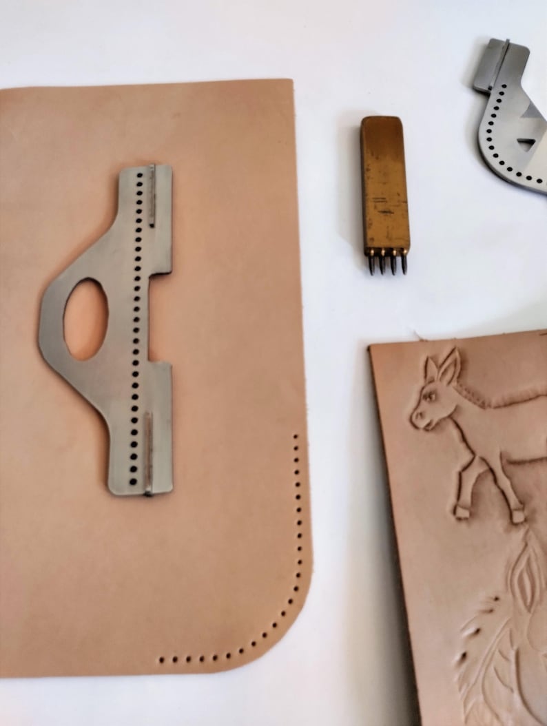 Peut inclure: Ensemble d'outils et de mat&eacute;riaux de maroquinerie. Comprend un guide de couture en m&eacute;tal, un poin&ccedil;on &agrave; plusieurs dents et une pi&egrave;ce de cuir avec un motif d'&acirc;ne. Les outils sont dispos&eacute;s sur une surface blanche, mettant en valeur le travail.