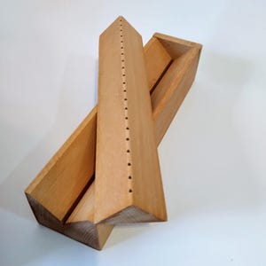May include: A wooden box with a triangular top and a series of holes along the top edge. The box is open and the inside is visible. The box is made of light wood.