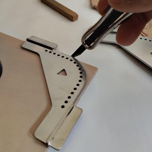Peut inclure: Un outil de travail du cuir en acier inoxydable avec un bord incurv&eacute; et des trous espac&eacute;s de mani&egrave;re uniforme est utilis&eacute; pour couper un morceau de cuir brun clair. Un petit outil en laiton et un couteau &agrave; couper le cuir sont &eacute;galement visibles.