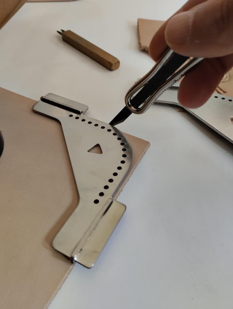 Peut inclure: Un outil de travail du cuir en acier inoxydable avec un bord incurv&eacute; et des trous espac&eacute;s de mani&egrave;re uniforme est utilis&eacute; pour couper un morceau de cuir brun clair. Un petit outil en laiton et un couteau &agrave; couper le cuir sont &eacute;galement visibles.