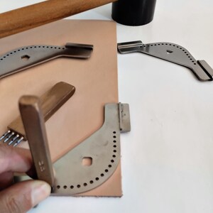 Peut inclure: Un ensemble d'outils de maroquinerie, comprenant un maillet en bois, un objet cylindrique noir et plusieurs outils d'angle en m&eacute;tal avec des trous espac&eacute;s. Une pi&egrave;ce de cuir beige est &eacute;galement pr&eacute;sente.