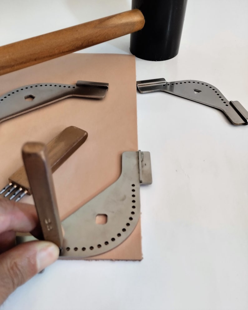 Peut inclure: Un ensemble d'outils de maroquinerie, comprenant un maillet en bois, un objet cylindrique noir et plusieurs outils d'angle en m&eacute;tal avec des trous espac&eacute;s. Une pi&egrave;ce de cuir beige est &eacute;galement pr&eacute;sente.