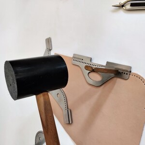 Peut inclure: Un maillet noir avec un manche en bois est pos&eacute; &agrave; c&ocirc;t&eacute; d'un morceau de cuir beige. Des outils en m&eacute;tal avec une s&eacute;rie de trous sont positionn&eacute;s pr&egrave;s du cuir, sugg&eacute;rant le travail du cuir. Une paire de pinces m&eacute;talliques est visible en arri&egrave;re-plan.