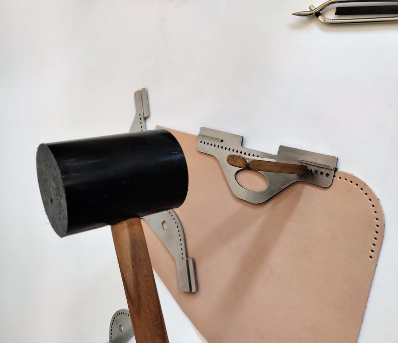 Peut inclure: Un maillet noir avec un manche en bois est pos&eacute; &agrave; c&ocirc;t&eacute; d'un morceau de cuir beige. Des outils en m&eacute;tal avec une s&eacute;rie de trous sont positionn&eacute;s pr&egrave;s du cuir, sugg&eacute;rant le travail du cuir. Une paire de pinces m&eacute;talliques est visible en arri&egrave;re-plan.