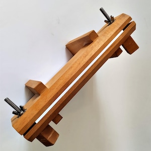 May include: A wooden leathercraft clamp with a natural finish. The clamp features a long, rectangular body with a central channel and adjustable wing nuts. The wood is light brown, and the clamp is designed for holding leather pieces.