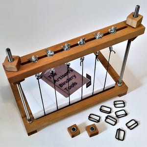 May include: A wooden bookbinding press with a book inside, five metal screws, and eight black metal buckles. The press is labeled "The Artisan's Bindery Tools".