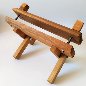 Può includere: Un supporto per telaio da ricamo in legno con finitura naturale. Il supporto presenta due barre orizzontali tenute insieme da aste filettate e dadi ad alette, sostenute da gambe angolate. La venatura del legno è visibile.