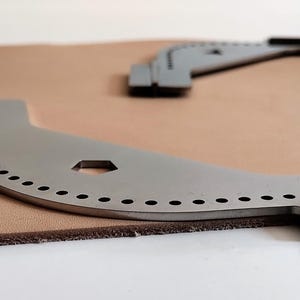 Peut inclure: Deux outils de travail du cuir argent&eacute;s, aux bords incurv&eacute;s et &agrave; multiples trous, sont pos&eacute;s sur un morceau de cuir beige. Un outil est au premier plan, l'autre &agrave; l'arri&egrave;re-plan. Les outils ont une extr&eacute;mit&eacute; plate et rectangulaire.