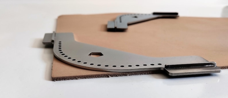 Peut inclure: Deux outils de travail du cuir argent&eacute;s, aux bords incurv&eacute;s et &agrave; multiples trous, sont pos&eacute;s sur un morceau de cuir beige. Un outil est au premier plan, l'autre &agrave; l'arri&egrave;re-plan. Les outils ont une extr&eacute;mit&eacute; plate et rectangulaire.