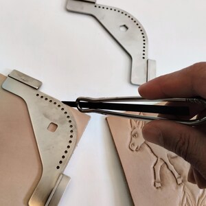 Peut inclure: Outils de travail du cuir en acier inoxydable. L'image montre un rainureur &agrave; coudre en m&eacute;tal et deux gabarits m&eacute;talliques avec des trous espac&eacute;s. Un morceau de cuir avec un motif d'&acirc;ne est &eacute;galement visible. Ces outils sont utilis&eacute;s pour le travail du cuir.