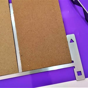 Op de afbeelding: Een rechthoekig stuk bruin vezelplaat bevestigd aan een zilverkleurige metalen beugel. De metalen beugel is voorzien van een platte horizontale rand, een verticaal uitsteeksel met een driehoekige uitsparing en een vierkant gat rechtsonder.