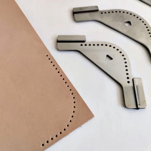Peut inclure: Gros plan sur un morceau de cuir beige avec un bord incurv&eacute;, ponctu&eacute; de trous espac&eacute;s. Trois outils de maroquinerie en m&eacute;tal sont positionn&eacute;s &agrave; proximit&eacute;. Les outils ont un design incurv&eacute; avec une s&eacute;rie de trous et une extr&eacute;mit&eacute; rectangulaire.