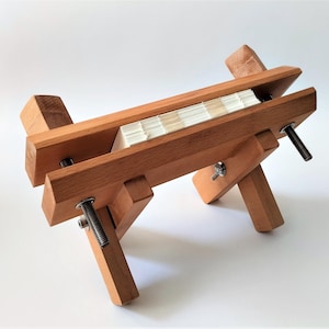 Op de afbeelding: Een houten boekbindpers met twee verstelbare klemmen en een stapel papier erin. De pers is gemaakt van lichtbruin hout en heeft een natuurlijke afwerking.