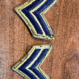 May include: Two chevron-shaped embroidered patches featuring a dark blue center stripe bordered by light yellow stitching. The patches have a distinct V-shape design with thick, textured thread work and a matching light yellow fabric border.