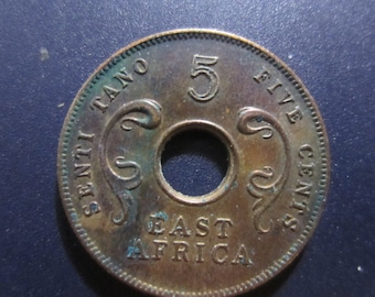 Könnte beinhalten: Eine kupferfarbene Münze mit einem Loch in der Mitte. Die Münze trägt die Aufschriften "Five Cents" und "East Africa". Die Münze trägt auch die Zahl "5" und das Wort "Tano" ein.