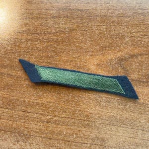 Peut inclure: Un écusson en tissu étroit en forme de parallélogramme, composé d'une base en feutrine bleu marine foncé avec une section rectangulaire centrale allongée de fil à broder vert métallique scintillant cousu en diagonale.