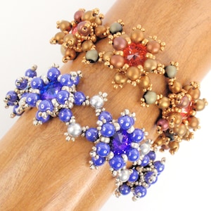 Könnte beinhalten: Drei Perlenarmbänder mit sternförmigen Designs. Das obere Armband ist gold und braun mit einem roten Strassstein in der Mitte. Das mittlere Armband ist blau mit einem violetten Strassstein in der Mitte. Das untere Armband ist blau mit einem silbernen Strassstein in der Mitte.