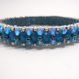 May include: A teal blue beaded bracelet with iridescent blue and silver beads. The bracelet is made with a woven pattern and has a delicate, feminine look.