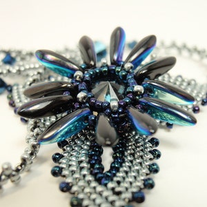 Puede incluir: Primer plano de un collar de cuentas plateadas y azules con una gran pieza central iridiscente negra y azul. La pieza central tiene forma de flor con múltiples pétalos.