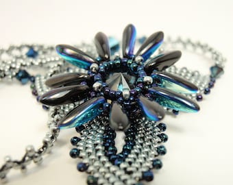 May include: A close-up of a silver and blue beaded necklace with a large, iridescent, black and blue centerpiece. The centerpiece is shaped like a flower with multiple petals.