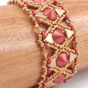 Puede incluir: Un brazalete de cuentas doradas y rosadas con un patrón geométrico. El brazalete está hecho con cuentas de semillas pequeñas y cuentas redondas más grandes.