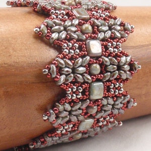 May include: A beaded bracelet with a geometric design featuring silver, gray, and red beads. The bracelet is made with a variety of bead shapes and sizes.