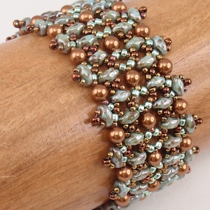 May include: A beaded bracelet with a woven pattern in shades of green, brown, and copper. The bracelet is made with small seed beads and larger round beads.