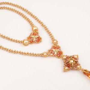 Könnte beinhalten: Eine goldene und pfirsichfarbene Perlenkette mit einem tropfenförmigen Kristallanhänger. Die Kette hat ein mehrschichtiges Design mit zwei Perlensträngen und einem dekorativen Mittelstück.