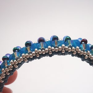 May include: A close-up of a beaded bracelet with a blue, silver, and black color scheme. The bracelet is made with small, round beads and has a slightly curved shape.