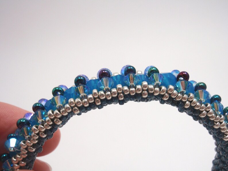 May include: A close-up of a beaded bracelet with a blue, silver, and black color scheme. The bracelet is made with small, round beads and has a slightly curved shape.