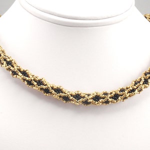 Op de afbeelding: Een kralenketting met een goud en zwart ontwerp. De ketting is gemaakt van kleine, ronde kralen die aan elkaar zijn geweven om een ​​gestructureerd, touwachtig uiterlijk te creëren. De gouden kralen worden afgewisseld met zwarte kralen.