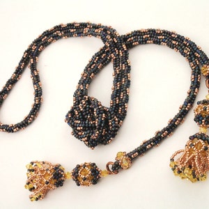 以下が含まれることがあります： ダークブルーと銅色の手作りビーズネックレス。 ネックレスは、ビーズのアクセントとタッセルのペンダントが付いた、ねじれたロープのデザインです。 ユニークなジュエリーです。