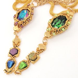 Op de afbeelding: Een gouden ketting met een hanger van groene, paarse en iriserende blauwe edelstenen. De hanger is gemaakt van kralen en heeft een geometrisch ontwerp.