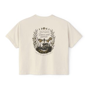 May include: An off-white t-shirt featuring a dark graphic on the back. The design includes a house, two cocktail glasses, a butterfly, and moon phases, with the text: BAD DECISIONS. GOOD COMPANY. Dance until sunrise. YOU PUT THE LIME IN THE COCONUT.