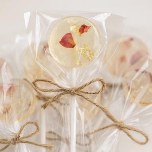 May include: Clear lollipops with embedded rose petals and gold flakes, individually wrapped in cellophane. Each lollipop is tied with a twine bow. The lollipops are on white sticks, creating a delicate and elegant treat.