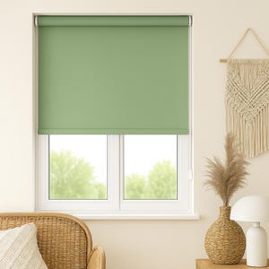 May include: A sage green roller shade is installed in a white window frame. The shade is pulled down, covering the window. The room is decorated with a woven vase, a macrame wall hanging, and a white lamp.