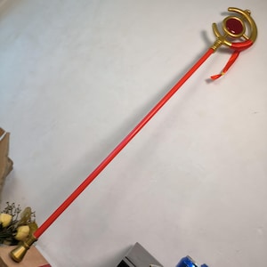 May include: A red staff with gold accents and a red gem. The top features a crescent moon design with a circular red gem in the center. A red ribbon is tied near the top. The staff has a gold base.