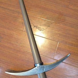 May include: A gray pickaxe with a long, cylindrical handle and a curved, pointed blade. The metal head has a sharp edge on one side and a pointed end on the other. The handle is made of a dark gray material.