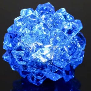 Könnte beinhalten: Ein Cluster aus facettierten, blauen Glasstücken, die an eine Kristallkugel erinnern. Das Glas hat eine reflektierende Qualität, wobei das Licht durch die Facetten scheint. Das Objekt befindet sich vor einem dunklen Hintergrund.