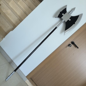 Könnte beinhalten: Eine große, zweischneidige Axt mit einem langen schwarzen Stiel. Der Axtkopf ist silber und schwarz, mit dekorativen Aussparungen. Die Axt ist wahrscheinlich ein Requisit oder ein Kostümteil.