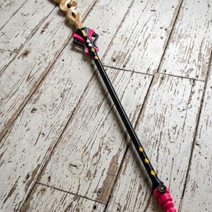 Op de afbeelding: Een fantasie-staf met een goudkleurige bovenkant, een zwarte schacht en roze accenten. De staf heeft een decoratief ontwerp met een puntig uiteinde. De staf staat op een verweerd, wit geschilderd houten oppervlak.