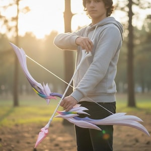 以下が含まれることがあります： ファンタジーをテーマにした弓矢セット。弓は白、ピンク、虹色のアクセントが付いた曲線的なデザインです。矢にはピンクの矢じりが付いています。弓はグレーのパーカーを着た人が持っています。