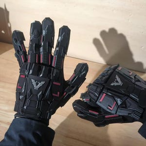 May include: Pair of black, articulated gloves with red accents and a silver emblem. The gloves have a futuristic, mechanical design, with segmented plates and a textured surface. The gloves are likely for cosplay or costume use.