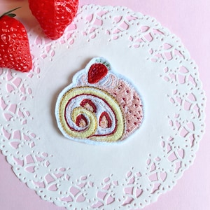 以下が含まれることがあります： 赤いイチゴがトッピングされたピンク色のスイスロールの刺繍パッチ。白いドイリーの上に配置されています。パッチには黄色と赤色のディテールがあります。背景はピンク色で、赤いイチゴが3つあります。